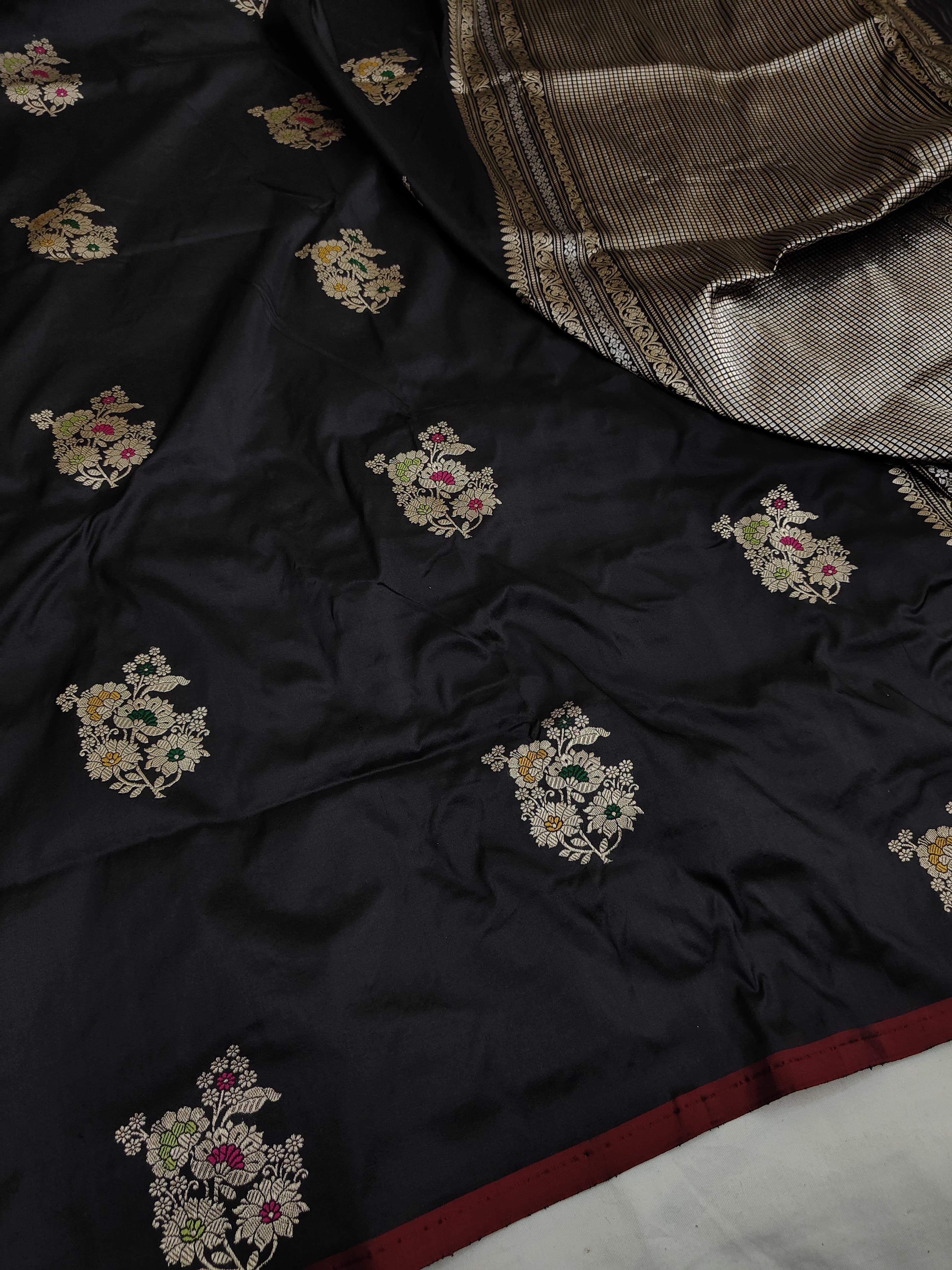 A close-up of a black silk saree or fabric with small floral motifs in gold, pink, and green, featuring a metallic gold and silver patterned border and a dark red edge.