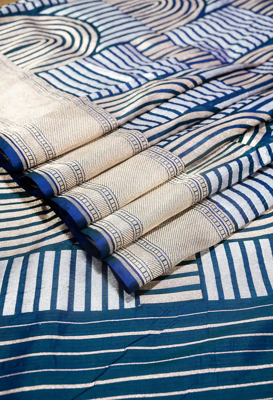 A close-up of a patterned fabric, likely a saree, featuring a cream-colored body with blue and white geometric stripes and a border, draped over a larger matching section.