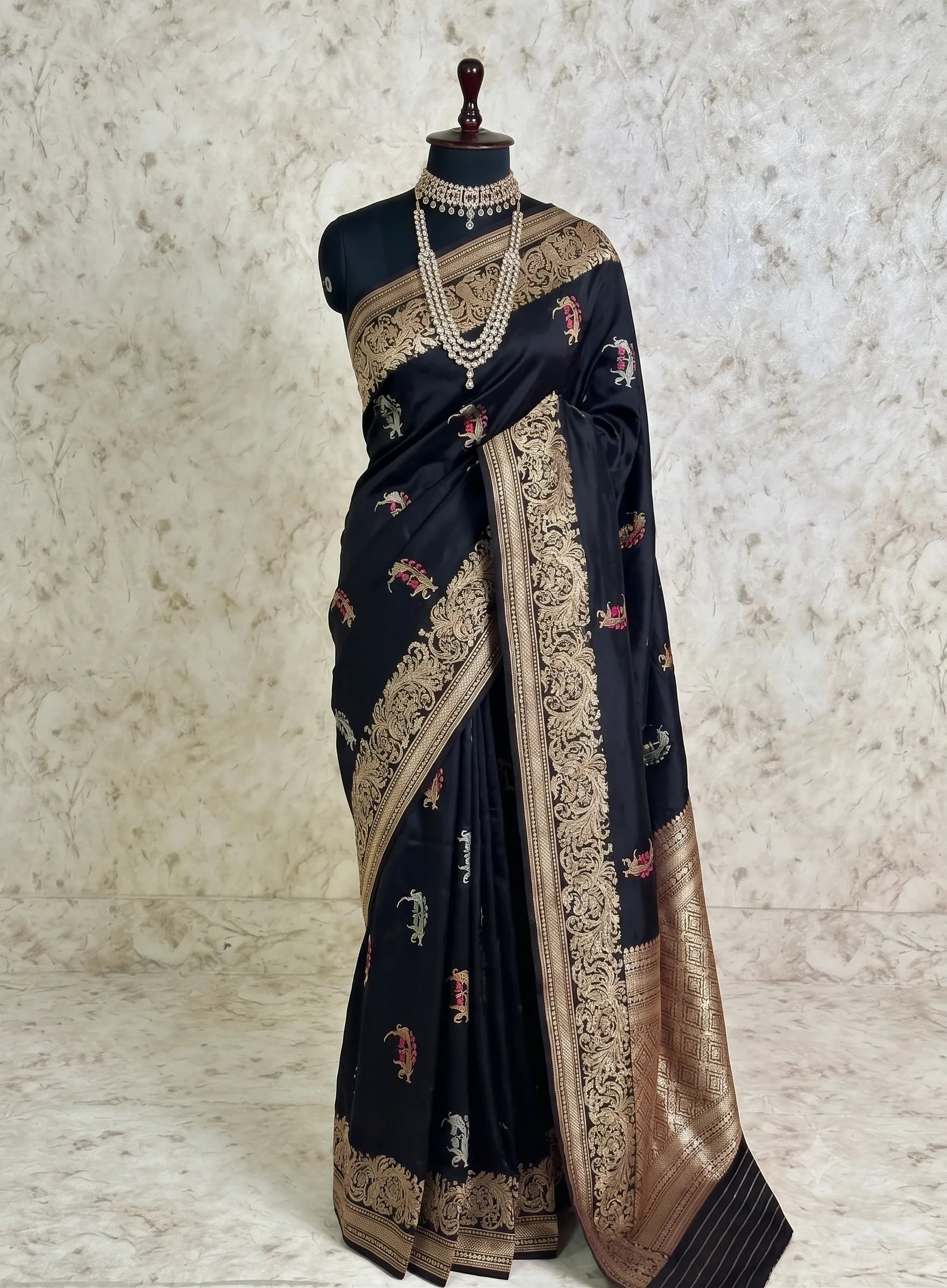 A black silk saree with intricate gold zari embroidery and small floral motifs, displayed on a black mannequin with a pearl necklace. The background is a textured beige wall and marble floor.