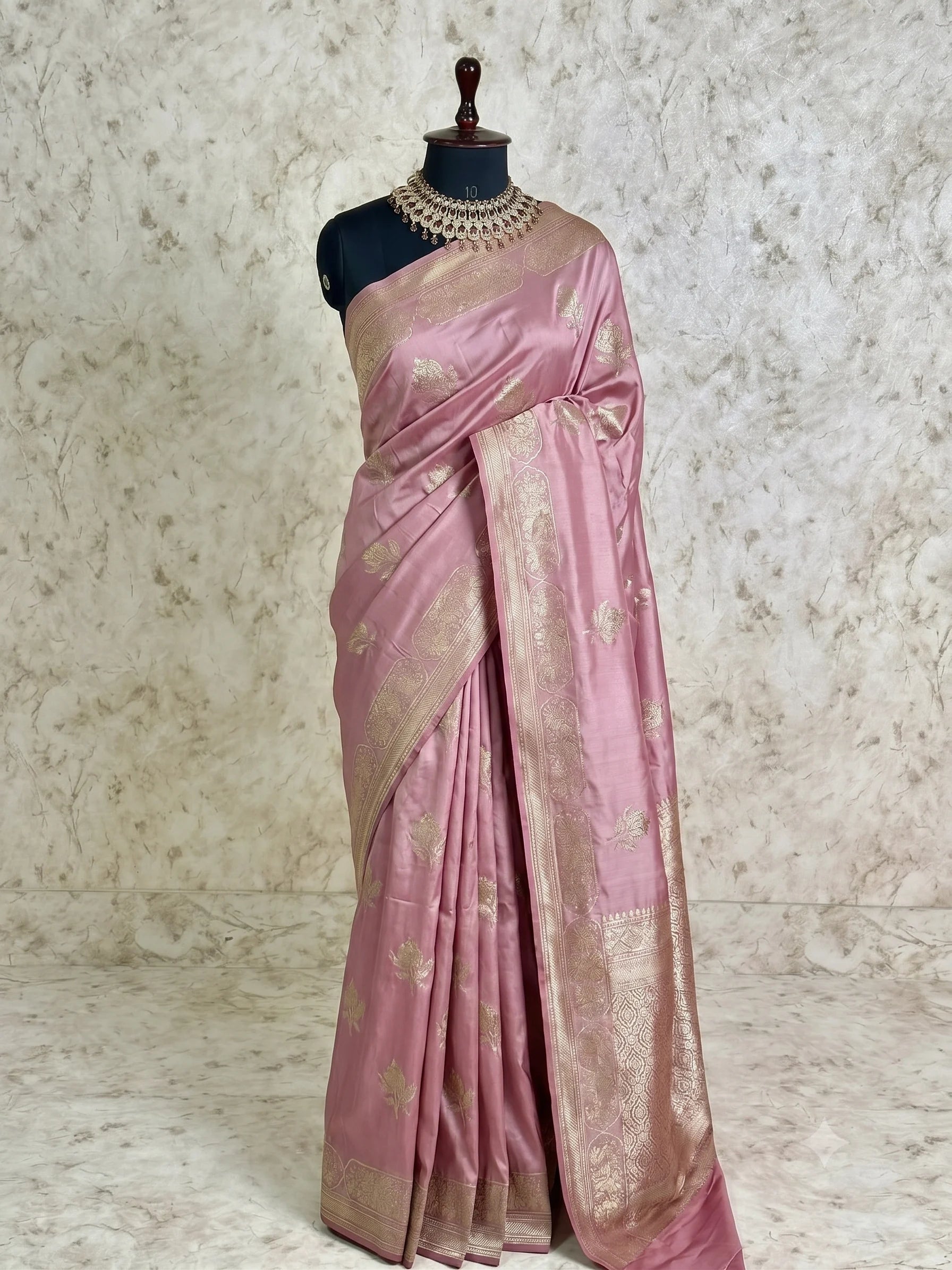 A pink silk saree with gold zari floral motifs and a wide gold border, draped on a black mannequin torso against a textured beige backdrop.