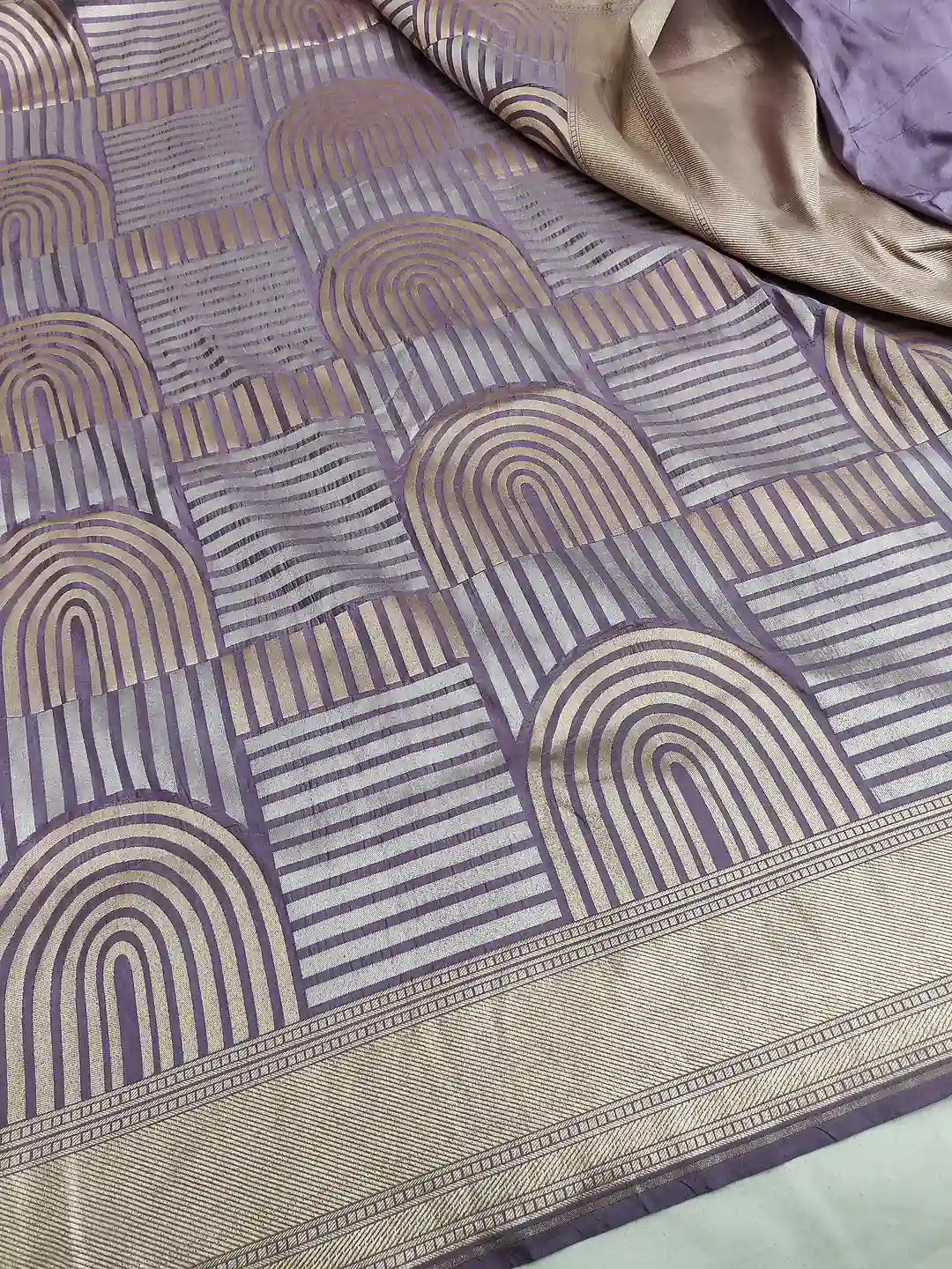 A close-up of a patterned fabric, likely a saree, featuring a geometric design with arches and stripes in lavender purple and beige. A gold-toned border is visible along the bottom edge, and another section of the fabric is draped in the top right corner.