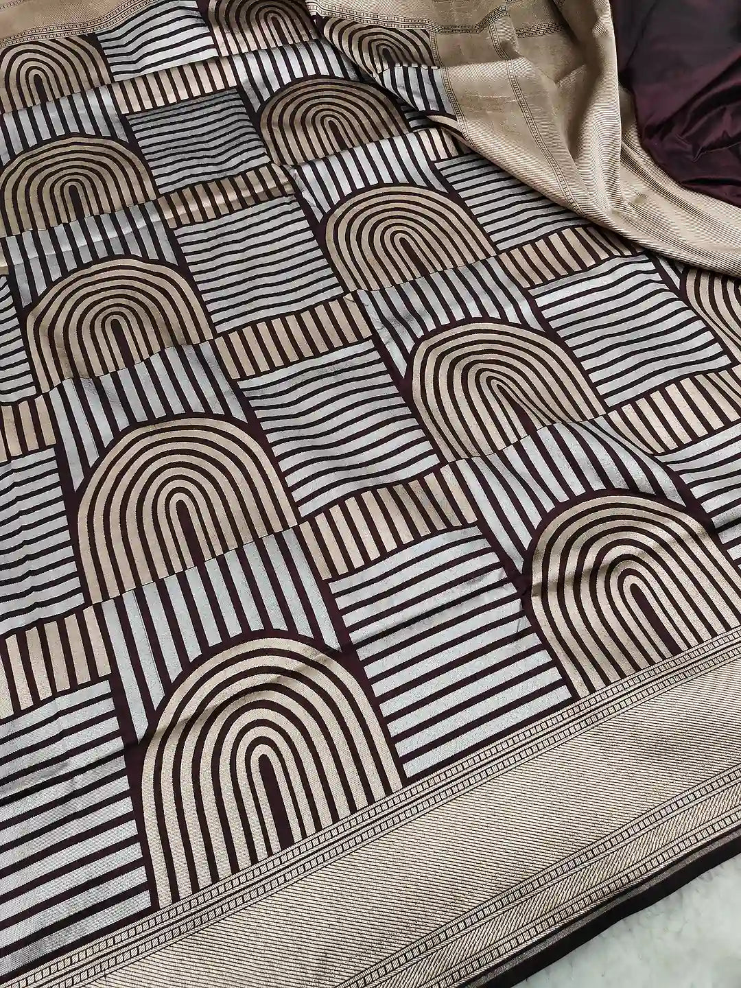 A close-up of a patterned fabric, likely a saree, featuring a geometric design with arches and stripes in beige, brown, and black. A beige border with a decorative woven pattern is visible along the bottom right, and a dark purple fabric is draped in the top right corner.