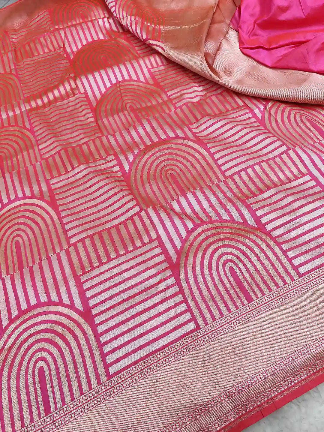 A close-up of a pink and white patterned fabric, likely a saree, featuring a geometric design with arches and stripes and a metallic border.