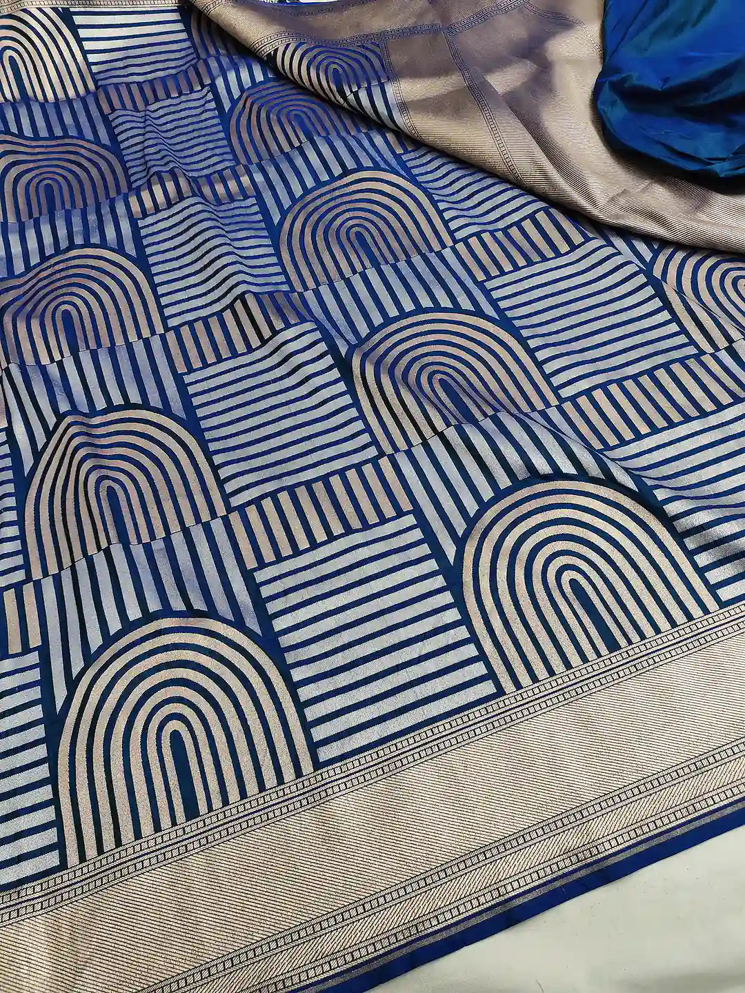 A close-up of a patterned fabric, likely a saree, featuring a blue and beige geometric design with arches and stripes, and a decorative border.