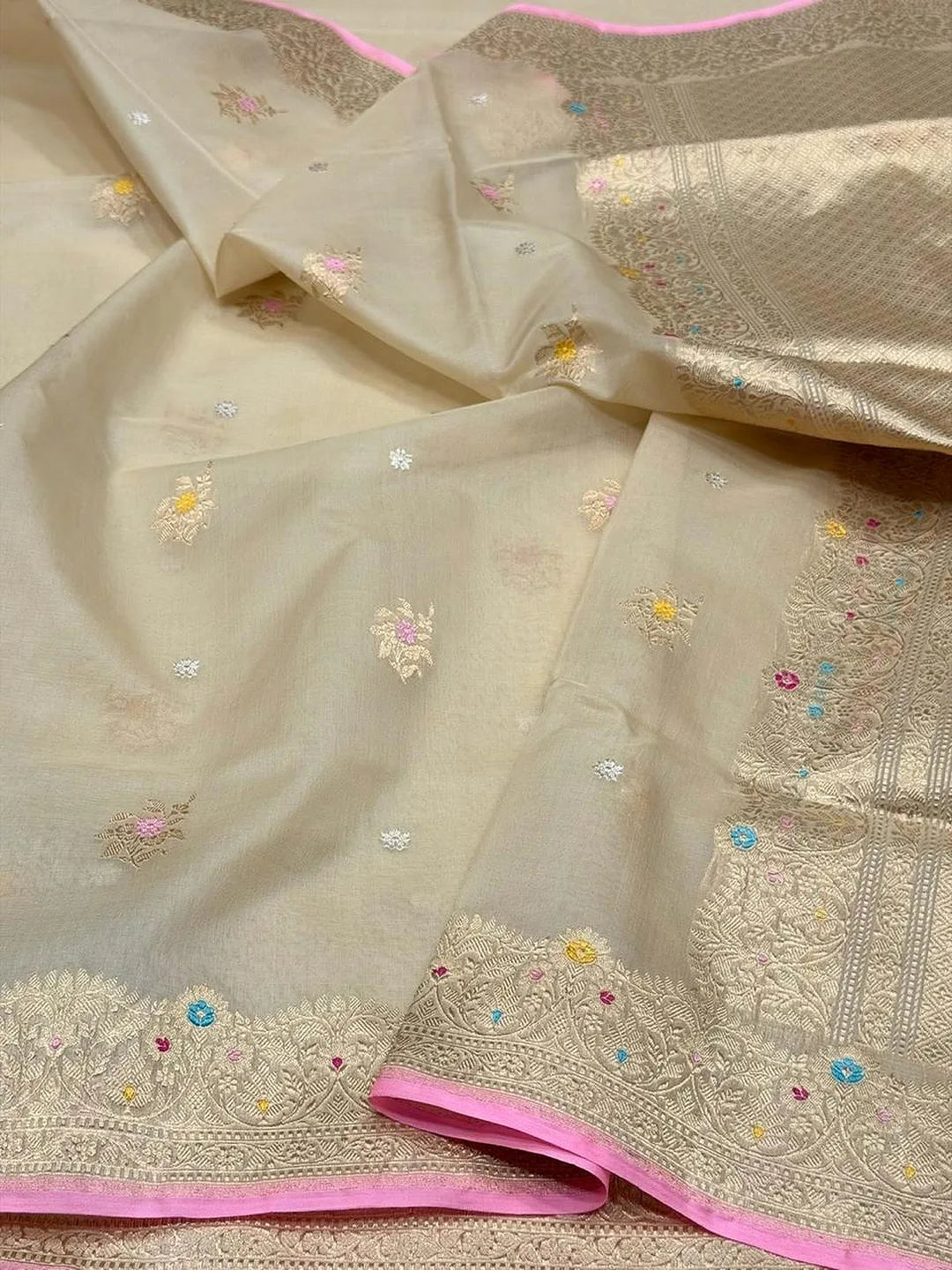 A close-up of a beige or cream-colored fabric with delicate floral embroidery, small colorful bead accents, and a bright pink border, shown draped in soft folds.